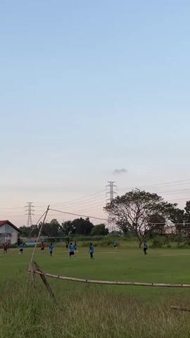 The video depicts a soccer field with several individuals engaged in a game. The sky is a pale blue, transitioning to a lighter hue near the horizon. Tall, lattice-like electricity towers are visible in the background, along with a cluster of trees. On the left side of the frame, a simple building with a red roof stands out. In the foreground, tall, dry grass and a makeshift wooden barrier frame the playing area. The players, dressed in blue and red attire, are spread across the field, some in motion, suggesting active gameplay.