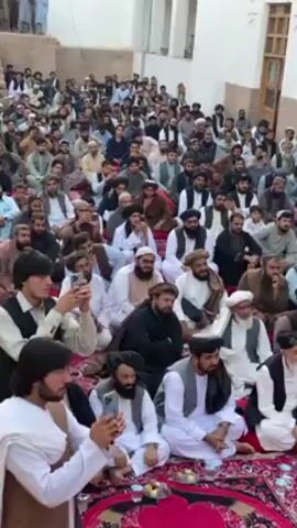 The image shows a large gathering of mostly men, seated on the ground on a red carpet. They are all dressed in traditional Afghan clothing, including shalwar kameez and vests. Many of the men have beards and wear turbans.

The scene appears to be an outdoor event, possibly a religious or social gathering, held in a courtyard with buildings visible in the background. The architecture suggests a traditional Middle Eastern or South Asian style. The lighting indicates it is daytime, likely morning or afternoon, with no strong shadows suggesting harsh midday sun. The weather appears to be pleasant, as people are seated outdoors without any visible signs of discomfort.

Some individuals in the foreground are looking at their phones, suggesting that the event is being documented or shared. There is a sense of community and shared experience among the attendees. The mood seems to be one of solemnity or respect, with most individuals facing the same direction, likely towards a speaker or focal point not entirely visible in the frame. The presence of food items on a platter in the foreground suggests a communal meal or offering as part of the event.

The location context of Kandahar, Afghanistan, further informs the interpretation of the scene, aligning with the traditional attire and architectural style. The image captures a moment of collective assembly in a cultural and potentially religious setting. No discernible text is visible in the image.