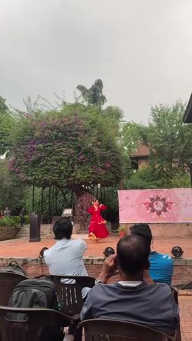 A woman in a red and gold dress is dancing on a brick stage in front of an audience. The stage is in an outdoor courtyard surrounded by greenery, including a large tree with purple flowers. Behind the stage is a bronze sculpture and a pink banner with a design on it. The audience members are seated in black chairs and are watching the performance. The sky is overcast. There are lights around the stage. One audience member is holding his phone to his ear. A black backpack is hanging on the back of one of the chairs.