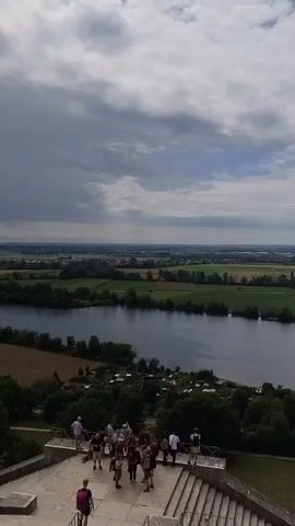 This is a vertical, high-angle, outdoor shot overlooking a wide river, with agricultural fields and a small village in the distance under a cloudy sky. In the foreground, a group of people are gathered on a concrete structure, possibly stairs or a platform, some walking and some standing. A large body of water, a river or lake, occupies the middle ground, with green trees lining its banks. Beyond the water, patchwork fields of varying shades of green and brown stretch towards the horizon, interspersed with more trees. Small clusters of buildings can be seen in the far distance. The overall scene depicts a serene landscape on a partly cloudy day.