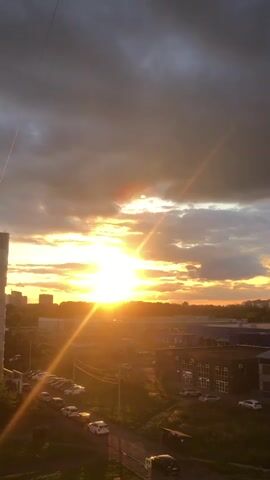 A time-lapse video shows a sunset over an urban landscape. The sun, a bright orb, is low on the horizon, casting long shadows and illuminating the sky with warm, golden light. Clouds, ranging from wispy white to dark, ominous gray, drift across the sky, creating a dynamic contrast with the sunlight. The cityscape below features various buildings, some appearing to be residential apartments and others larger commercial structures. A street with parked cars is visible, with a few vehicles in motion. The overall atmosphere is serene, with the changing colors of the sky reflecting the natural beauty of a sunset transitioning into twilight.