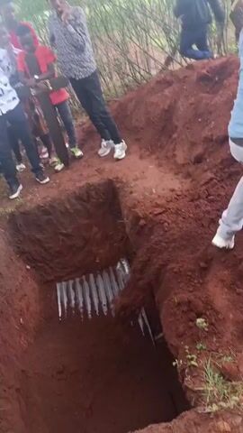 The video depicts a group of people gathered around a freshly dug rectangular hole in the ground, presumably a grave. The soil is reddish-brown. A man wearing a light blue shirt, grey jeans, white sneakers, and a yellow hat is seen shoveling dirt into the grave. Inside the grave, a corrugated metal sheet is visible. Other people are standing around the edge of the grave, observing. The environment appears to be a grassy area with some trees and bushes. A yellow flower lies at the edge of the grave.