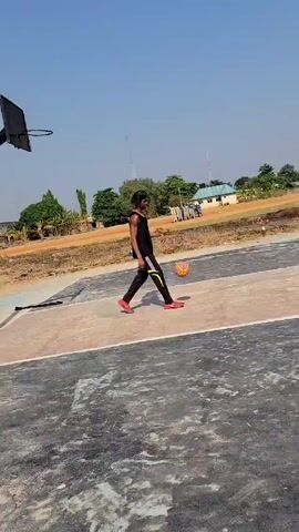 A young man practices basketball on an outdoor court under a bright blue sky. He is wearing a black tank top, black pants with yellow stripes, and red shoes. He dribbles a yellow basketball, moving around a painted circle on the gray and beige court. In the background, there is a yellow truck, trees, and a building. Another person walks across the background carrying something on their head.