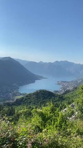 The video pans across a panoramic view of a bay surrounded by steep, tree-covered mountains under a clear blue sky. In the foreground, lush green foliage and some flowering plants are visible, with hints of red and orange blooms. As the camera moves, the bay opens up, revealing a body of turquoise water with a few small boats. Along the shore of the bay, a town with red-tiled roofs and buildings is nestled against the mountainside. The terrain is rugged and mountainous, with the town appearing to be built on the lower slopes. The overall impression is of a scenic and tranquil coastal landscape.