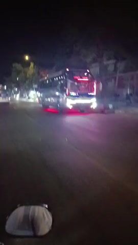 A motorcycle is traveling on a street at night. The motorcycle is moving forward and the camera is mounted on it, giving a first-person perspective. In the video, there are other vehicles on the road, including a large bus with red underglow lights and several cars. The street is lined with trees and buildings, some of which have illuminated signs. The road appears to be wet. The sky is dark, indicating it is nighttime. There are streetlights casting a yellow glow on the road.
