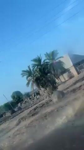 The video shows a view from a moving vehicle, likely a car or bus, traveling past rural landscape.  The scene features a clear, bright blue sky with power lines running across the top of the frame. The foreground consists of dry, scrubby vegetation and a dirt road, while in the background, there are sparse buildings or houses with corrugated metal roofs.  One structure is painted white and red.  The overall impression is of a dry, dusty environment.  There appears to be at least one person visible near one of the buildings.