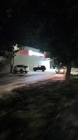 This is a nighttime video of a parking lot in front of a store. The store has a lit sign that says "ПОБЕДА". There are three cars in the parking lot. One is a white SUV parked on the left. Another is a black SUV parked in the middle. A third car is driving towards the viewer on the right. The black SUV has its hazard lights on. There is a tree on the right side of the frame. The ground is paved.