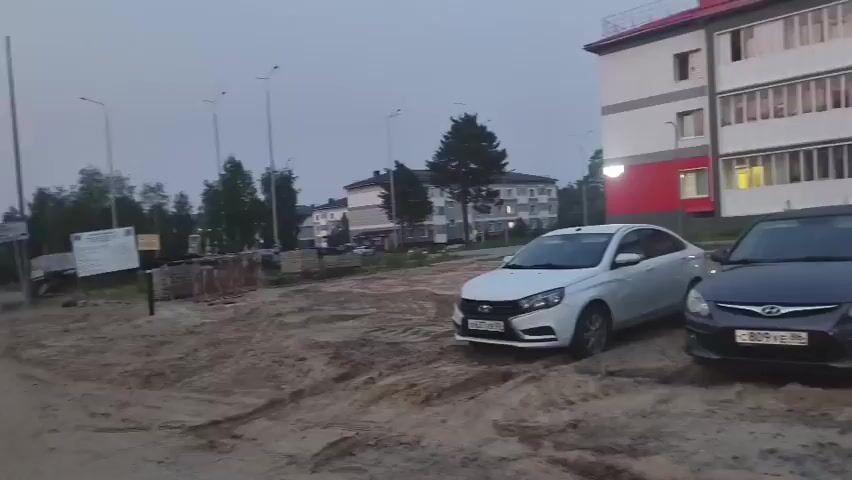 The video shows a construction site with several parked cars and an excavator. The buildings in the background appear to be apartment buildings. The ground is covered in dirt and sand, indicating that construction is ongoing. There are no people visible in the foreground, but a few people can be seen walking in the background. The sky is overcast, suggesting it is either early morning or late evening. The excavator is a large yellow machine with tracks. The cars are of various colors and models. The apartment buildings are modern in design, with red roofs and white and red facades. The overall scene suggests a developing residential area.