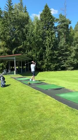 A person is seen practicing golf at a driving range. The person is wearing a t-shirt and shorts and is swinging a golf club. There is a golf bag next to them. The driving range has several mats for players to stand on, and a covered area in the background. The scene is set in a grassy area surrounded by trees under a clear blue sky. Another person is standing on the grass to the left of the frame.