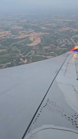 The video shows an aerial view from an airplane window, looking down at a landscape. The right wing of the airplane is prominently visible in the foreground, taking up the lower half of the frame. The wing is light gray and shows various rivets and seams, typical of aircraft construction. The tail fin of the airplane is visible at the end of the wing, displaying a colorful logo in red, yellow, and blue. Below the wing, a patchwork of agricultural fields and green terrain stretches out towards the horizon. Small clusters of buildings, indicating villages or towns, are scattered throughout the landscape. The sky appears hazy, suggesting either distance or atmospheric conditions. The camera is stationary, and the view is stable, implying the airplane is in steady flight. No people are visible.