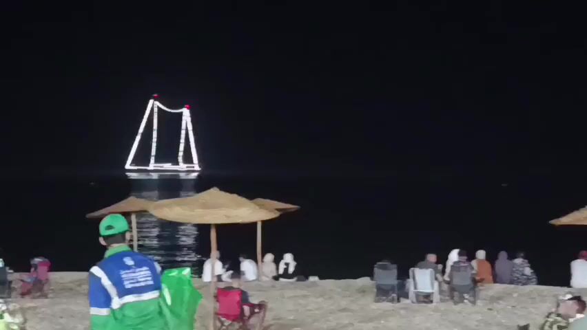 The video shows a nighttime beach scene. A large, illuminated structure resembling an A-frame or a simplified Eiffel Tower stands in the dark water. On the sandy beach, several groups of people are gathered, sitting under straw umbrellas or on beach chairs. A person in a blue and green uniform, possibly a lifeguard or event staff, is visible in the foreground, facing away from the camera. The overall atmosphere appears to be relaxed and social, with people enjoying the evening by the water.
