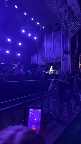 A concert is underway, with a band performing on a brightly lit stage under a deep purple hue. The stage is equipped with a large sound system and a backdrop of draped fabric. A crowd of people stands in front of the stage, many holding up their phones to record or take pictures. One person in the foreground is filming the concert with a smartphone. The lighting is dramatic, creating a vibrant and energetic atmosphere. The overall scene captures the excitement of a live musical performance.