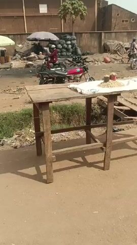 The scene is an outdoor street view during the day. The ground is a light brown dirt or pavement. In the foreground, a simple, wooden table stands, partially covered with a white cloth. On the table, there's a scattering of what appear to be peanuts. Behind the table, there's a strip of vegetation or grass. Further back, several motorcycles are parked. Some are ridden by people. In the background, several market stalls can be seen with people standing around them. There are also large, dark bags piled in one area. Buildings form the backdrop, with a tall tree behind the pile of bags. A person walks on the left side of the frame, carrying a large object on their head. The scene suggests a market area in a developing country.