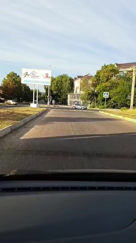This video is captured from the interior of a vehicle, looking forward. The vehicle is on a paved road, appearing to be approaching a crosswalk. To the left of the road, there is a sidewalk with a grassy verge and trees. A large billboard is visible on the left, displaying text and images. Further down the road on the left, a yellow car is parked. On the right side of the road, there are buildings and more trees. A silver car is moving away from the camera on the right side of the road. A pedestrian crossing sign is visible on the right. The sky is blue with scattered clouds, and the lighting suggests it is daytime, possibly morning or afternoon. The video shows movement as the vehicle drives forward and the scene changes slightly.