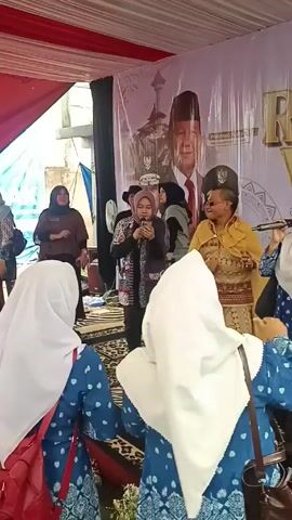 A group of women are gathered under a tent singing karaoke with a microphone, with a banner in the background featuring a portrait of Prabowo Subianto, an Indonesian politician and former general. Some of the women are wearing hijabs. They appear to be enjoying themselves, some dancing along to the music. The tent is decorated with red and white fabrics. The overall atmosphere seems festive.