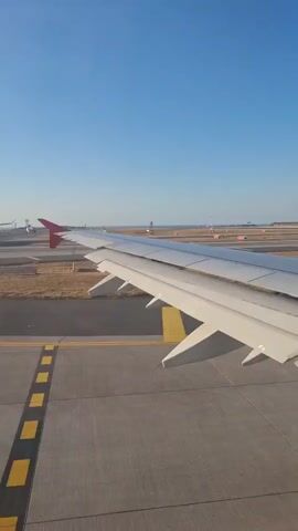 The video shows a view from an airplane window as it taxis on a runway. The airplane's wing, with its red-tipped end, is prominently visible in the foreground. The wing is extended and appears to be in good condition. The runway surface is visible, with yellow and white markings indicating the taxiway. In the background, other airplanes and airport infrastructure can be seen, including lights and signs. The sky is clear and blue, suggesting good weather conditions. The airplane appears to be moving slowly, as it is still on the ground and has not yet taken off.