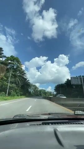 The video shows a first-person perspective from inside a vehicle, looking out at a road on a sunny day with a blue sky and white clouds. The road appears to be a two-lane highway with a concrete barrier on the right side. Trees and vegetation line both sides of the road. In the distance, the road curves to the left, and there are signs indicating curves ahead. The dashboard of the vehicle is visible in the foreground. The weather appears to be clear and warm, suggesting it is daytime. No people are visible within the frame.