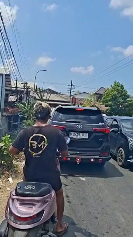 A person on a scooter is riding on a road. The person is wearing a black t-shirt with a logo on the back. The scooter is pink. There is a black SUV in front of the scooter, and other vehicles are visible. The sky is blue with white clouds. The road is lined with buildings and vegetation.