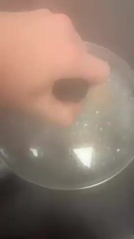 The video shows three eggs boiling in a pot of water. A hand removes the lid of the pot to reveal the boiling water and eggs. The pot is on a black stovetop. The video is shot from above. Steam rises from the pot.