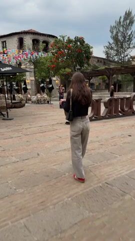 The video shows a woman walking across a brick-paved courtyard. She is wearing light grey jeans, a black top, and red sneakers, and carrying a black purse. The courtyard is decorated with colorful flags strung between buildings. There are several outdoor dining areas with tables and chairs, some covered by black umbrellas. Other people can be seen in the background, including someone taking a photo. Some buildings are visible with lights strung between them. The sky is overcast.