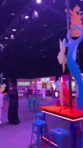 The video showcases a large, brightly lit event space, seemingly indoors, with a high ceiling and dark backdrop. The lighting is predominantly purple and blue, creating a vibrant atmosphere. There are several people, some wearing traditional Middle Eastern clothing, milling around the space. A tall, whimsical sculpture with abstract shapes and colors stands prominently. Various booths and structures are set up, including one labeled "THE UPSIDE," suggesting a theme or event concept. There are also gaming machines and retro-style displays. People are walking, observing, and engaging with the various attractions in the space.