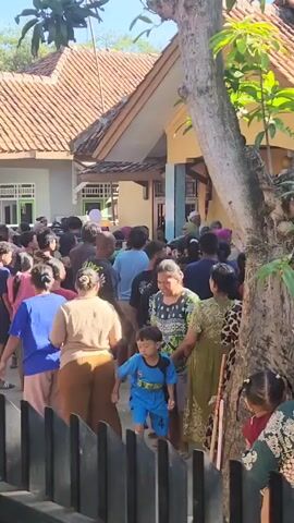 From behind a black fence, a crowd of people stands outside two houses, their hands raised in the air. One house is yellow with a brown tiled roof, the other is light grey with a brown tiled roof. Most people are facing the houses. There is a tree on the right and other greenery is visible. There are people of different ages and gender, mostly wearing casual clothing. A few children are visible in the front of the crowd.