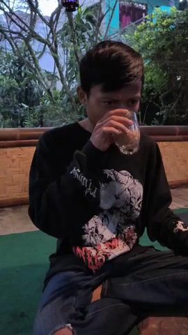 A young man with dark hair drinks from a glass and then sticks his tongue out and screams, then smiles and turns to his right. He is wearing a black long-sleeved shirt with a large graphic design and blue jeans. He is sitting cross-legged on a green surface in what appears to be an outdoor patio area, with trees, a bamboo railing, and a hanging lantern visible.  The overall environment is dimly lit.
