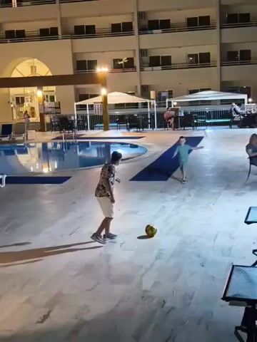 In this video, two young boys are playing with a soccer ball on a tiled patio. They appear to be at a resort, as a swimming pool is visible in the background, as well as lounge chairs and umbrellas. The hotel building behind the patio is lit up, indicating it is nighttime. The lighting suggests it might be evening or nighttime. One boy is wearing a floral-patterned shirt and white shorts, while the other is wearing a green t-shirt and shorts. They are kicking the ball back and forth. 

There are several tables and chairs set up on the patio, and a few adults are seated at some of them, observing the boys or engaged in conversation. The overall mood seems relaxed and casual, with the sounds of children playing and adult chatter filling the air. The scene appears to be set in a warm climate, judging by the attire of the people and the outdoor setting. The presence of palm trees or other tropical vegetation is not clearly visible, but the general ambiance suggests a holiday destination. The hotel architecture is typical of resorts, with balconies and large windows. The tiled patio floor reflects the artificial lights, creating a bright and inviting atmosphere.