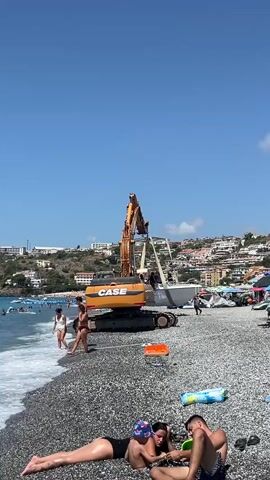 An orange excavator with "CASE" written on its side is positioned on a pebble beach next to the ocean. The excavator's boom is extended, lifting a white boat with a canopy. Several people are visible on the beach, some in swimwear. In the foreground, two individuals are lying on the beach, one in a black swimsuit and the other in swim trunks. The ocean is calm with small waves lapping at the shore. In the background, a town is built on a hillside overlooking the water. The sky is clear and blue.