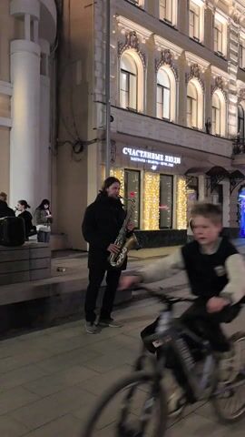 The video shows a male street musician playing a saxophone on a city street at night. He is wearing a black jacket and dark pants. A black bag with a white label displaying a phone number lies on the ground near his feet. Behind him, a building is visible with a sign in Russian that reads "Счастливые люди," which translates to "Happy People." Lights are strung around the storefront. Several people are standing and walking nearby, and a portable speaker is positioned to the left of the musician. The street is paved with tiles, and the overall atmosphere is urban and illuminated by streetlights and building lights.