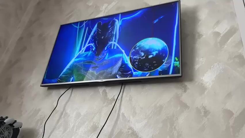 The video shows a television screen mounted on a textured wall. The television displays various scenes. Initially, a person in a futuristic suit with glowing blue accents is shown with a planet-like object to their right. Then, the scene changes to show two men, one in a suit and the other in a brown jacket and hat, engaged in what appears to be a confrontation or dialogue. The scene reverts to the person in the futuristic suit, with more emphasis on the blue and green lighting. Two black cables are visible hanging from the bottom of the television. A portion of a white door frame is visible on the right side of the screen.
