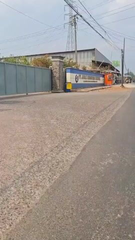 The video shows a road with a paved surface and a shoulder. On the left side of the road, there is a building with a sign that reads "PT JALE INVESTASI TEKNIK". Adjacent to the sign, there is a small orange kiosk. Further down the road, there are several small stalls and shops. The sky is overcast. On the right side of the road, there is a white fence with a decorative pattern and some trees behind it. A few more stalls and shops are visible along this side of the road as well. Power lines are visible overhead.