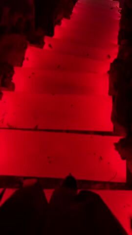 A person walks down a set of stairs. The stairs are illuminated by changing colored lights, transitioning from red to green. The walls adjacent to the stairs are rough and natural. In the background, there are hints of a dark environment with small, scattered blue lights, resembling stars or tiny glowing objects.