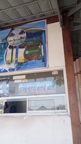 A video displays a ticket booth at an attraction. A large banner above the booth advertises the attraction, "Miana Saka Alas," with images of a park and a large decorative ball. Below the banner, a window displays pricing information for weekdays and weekends. A QR code is also visible. Inside the booth, a person wearing a white head covering and a dark top is seen interacting with someone outside the window. The booth has multiple windows, and one window has a sign that reads "BUKA OPEN". The environment appears to be outdoors, with a roof overhead.