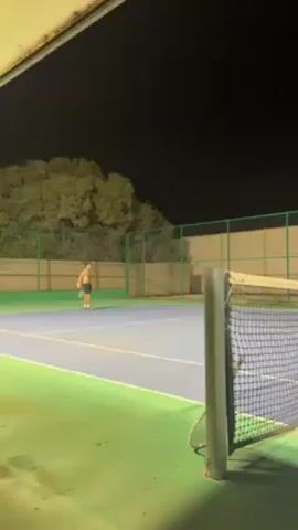 The video shows a man playing tennis on an outdoor court at night. The court is a combination of blue and green surfaces, surrounded by a green fence and a white wall. The court is lit by artificial lights. The tennis net is visible in the foreground, partially obscuring the view of the court. The man is shirtless and is striking a tennis ball with a racquet. The background includes trees in the distance under a dark night sky.