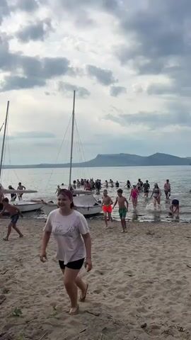 This video captures a group of people enjoying a beach setting. In the foreground, a young person wearing a white t-shirt and black shorts is running. Behind them, several other people are visible on the sand. To the left, two sailboats are partially submerged in the water, with more people gathered near them and in the water. The background shows a body of water, a distant shoreline with trees, and a cloudy sky. The scene appears to be a recreational gathering with activities taking place on the beach and in the water. The overall atmosphere suggests a casual and lively environment.