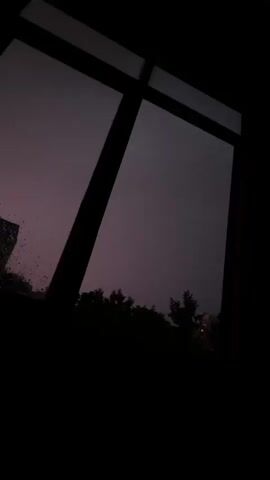 The video shows a window frame with a dark sky visible outside. There are trees silhouetted against the sky. A faint flash of lightning is visible in the distance. The scene appears to be during a storm at night.