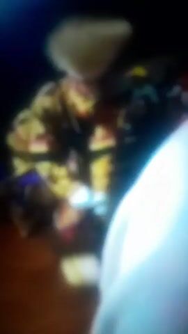 The video shows a gathering of people, likely outdoors at night, with a person in a patterned yellow and brown garment standing near the center. The image is blurry and details are indistinct, but a few individuals can be partially seen in the background. There is a rhythmic noise accompanying the scene, possibly music or chanting.