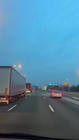 The video shows a forward-facing view from a vehicle moving along a multi-lane highway. The sky is overcast with a bluish tint, suggesting early morning or late evening. Several vehicles are present on the road, including multiple large semi-trucks and passenger cars. The camera car is in the rightmost lane. Highway signs are visible overhead indicating various destinations such as Arnhem, Nijmegen, and Rotterdam. Streetlights illuminate the roadway. The vehicle is moving, evidenced by the changing perspective relative to other vehicles and road markings.