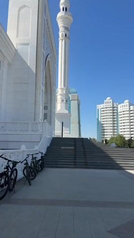 The video shows a wide shot of the exterior of a mosque with a tall minaret. The mosque is white with intricate blue and white decorative elements. In the foreground, to the left, are several bicycles parked along a low railing. A wide set of grey stairs leads up to the mosque entrance. A tall, modern blue glass building stands behind the minaret, and a white multi-story building is visible further to the right. The sky is clear and blue. A person is visible walking up the stairs toward the mosque.