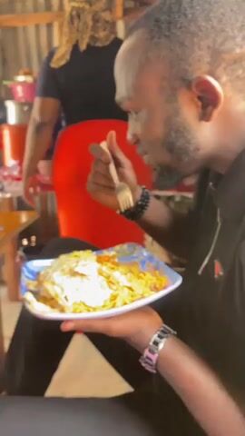 A man, with short dark hair and a beard, is holding a plate of food with his left hand and eating it with a fork in his right hand. He is wearing a black polo shirt and a wristwatch with a pink band on his left wrist. In the background, another person with blonde hair and wearing a red skirt is visible. The environment appears to be rustic, with corrugated metal walls.