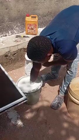 A man with dark curly hair, wearing a navy blue long-sleeved shirt and light-colored jeans, is seen bent over a bucket, mixing a white substance with his hand. He is surrounded by other containers, including a yellow jerrycan and another bucket filled with a tan liquid. The ground appears to be a mix of dirt and scattered white powder. A white-framed object with a dark center stands to his left, leaning against something out of frame. The background consists of a concrete wall. The scene appears to be outdoors, with strong sunlight casting shadows on the ground.