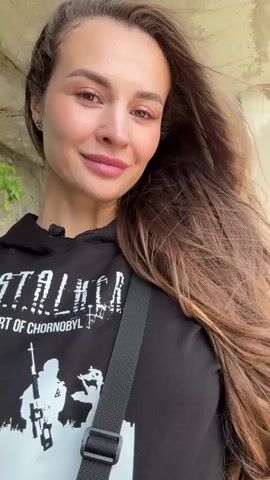 A young woman with long brown hair is filming a selfie video. She is wearing a black hoodie with a graphic design that says "STALKER HEART OF CHORNOBYL." She is smiling and looking at the camera. The background appears to be an abandoned building with concrete walls and some greenery.