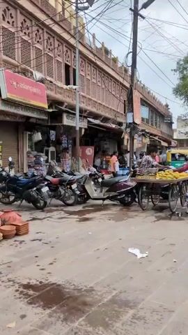 The video begins with a street scene in what appears to be India. A food cart selling corn on the cob is visible, with several people standing around it. Motorbikes and auto-rickshaws are parked or moving on the street. Buildings with ornate architecture line the street, some with shops and signs. The sky is overcast. The camera pans to the right, showing more of the street and shops. It then focuses on a white car parked in front of a building with an arched doorway and an air conditioning unit mounted on the exterior wall. The video ends with a close-up of a man in a pink shirt walking away from the camera, carrying a grocery bag. He is walking into an alleyway.