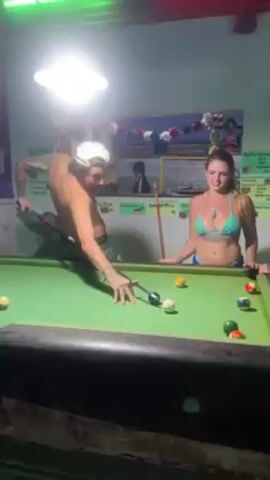 The video shows three people playing pool in a bar-like setting. A shirtless man with tattoos and a backwards cap is lining up a shot on a pool table covered in colorful pool balls. Two women in swimwear are standing on the right side of the pool table, watching the game. One is wearing a blue-green bikini and has tattoos, while the other has a short haircut and is wearing a black cut-out top. The woman in the blue-green bikini exclaims and is excited as the man shoots. The walls behind the pool table are decorated with posters and menus listing various food items like "Pad Thai" and "Green Curry." Overhead, there is bright lighting and some festive decorations.