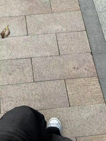 A person wearing black jeans and white sneakers is standing on a paved walkway. The walkway is made of rectangular light brown pavers. There is a gray concrete border along the right side of the walkway. A dead leaf is visible on the left side of the frame. The camera is positioned directly above the person's feet, looking down. The person does not appear to be moving.