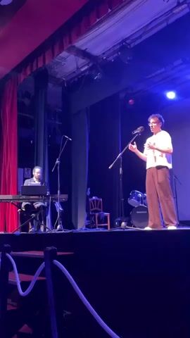 A man stands on a stage speaking into a microphone, gesturing with his hands. He is wearing glasses, a white t-shirt, and brown pants. To his left, another person sits at a keyboard. A drum set is visible behind the speaker. Red curtains hang on the left side of the stage, and blue lighting illuminates the scene. A dark railing is in the foreground.