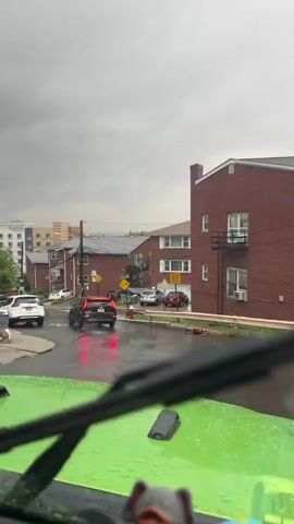 The video shows a view from inside a bright green vehicle looking out at a street scene during a rainy day. The road is wet and reflects the overcast sky. Several cars are driving on the street, including a white SUV, a black SUV with its brake lights on, and a red car further down the road. In the background, there are multi-story buildings, some appearing to be apartment complexes with modern architecture and others that look like older brick houses. Trees and utility poles line the street.

There are two yellow diamond-shaped road signs visible, one with an arrow indicating a curve and the other with the word "SLOW." On the wet pavement, the word "SLOW" is painted in large white letters. A red fire hydrant is also visible on the side of the road. The sky is heavily clouded, suggesting a stormy or heavily overcast day. The time of day appears to be daytime, likely afternoon, given the lighting. The presence of rain and the cautious driving suggest an event of driving in wet weather. The overall mood is neutral, focusing on the visual aspects of the street and traffic. The license plate of the white SUV has visible text, but it is partially obscured.