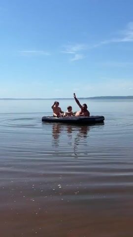 A man and two young children wave from an inflatable raft on a calm body of water under a bright blue sky. The adult is lying down in the raft. The older child is seated, and the youngest child is between the two. There are very faint ripples on the water surface and the reflection of the group on the water. The horizon line shows what appears to be land.