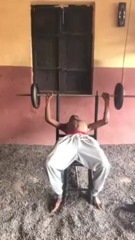 The video shows a slender, shirtless man with light-colored baggy pants lying on a weight bench and lifting a barbell with weights on each end. He is outdoors, on a gravel surface. Behind him is a wall painted both yellow and red, with a dark door. There is music in the background.